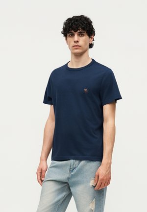 Jeune homme aux cheveux bouclés et foncés, portant un t-shirt bleu marine et un jean bleu clair déchiré, debout devant un fond blanc uni.