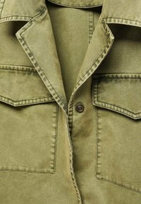 Detailní záběr na olivově zelenou košili s knoflíky, která má dvě náprsní kapsy a precizní prošívání kolem límce a švů.