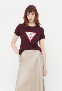 Mujer con cabello rojo viste una camiseta de logo de Guess en burdeos y una falda de tiro alto beige, de pie contra un fondo blanco liso.