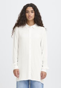 Sieviete ar gariem cirtainiem matiem valkā pārāk lielu baltu pogājamu kreklu un gaiši zilas džinsas, stāvot pret vienkāršu baltu fonu.