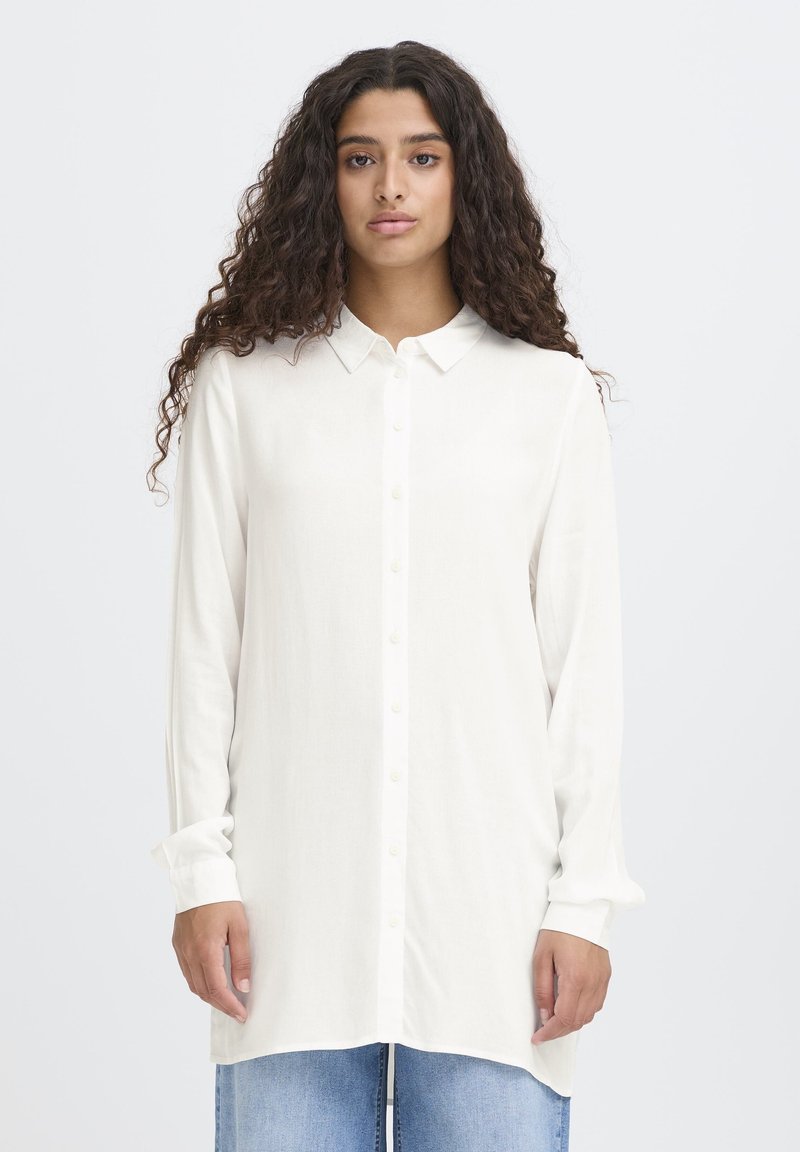 Sieviete ar gariem cirtainiem matiem valkā pārāk lielu baltu pogājamu kreklu un gaiši zilas džinsas, stāvot pret vienkāršu baltu fonu.