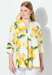 Blusa bianca con stampa di limoni e foglie, scollatura a V e maniche a tre quarti, realizzata in tessuto leggero. Bracciale dorato visibile.