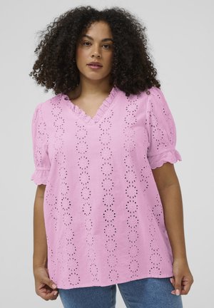 Frau mit lockigem Haar, die eine rosa Bluse mit Lochmuster, Rüschensaum am V-Ausschnitt und an den Ärmeln trägt, kombiniert mit blauen Jeans, steht vor einem schlichten Hintergrund.