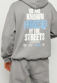 Conjunto de sudadera y pantalones deportivos grises con capucha. Diseño con capucha y texto en blanco y azul en la parte trasera. El material parece suave con puños y cintura acanalados.