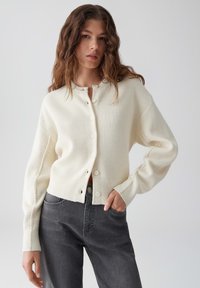 Witte gebreide cardigan met knopen aan de voorkant, verlaagde schouders en geribde manchetten; gedragen over donkergrijze jeans. Gladde textuur, casual ontwerp.