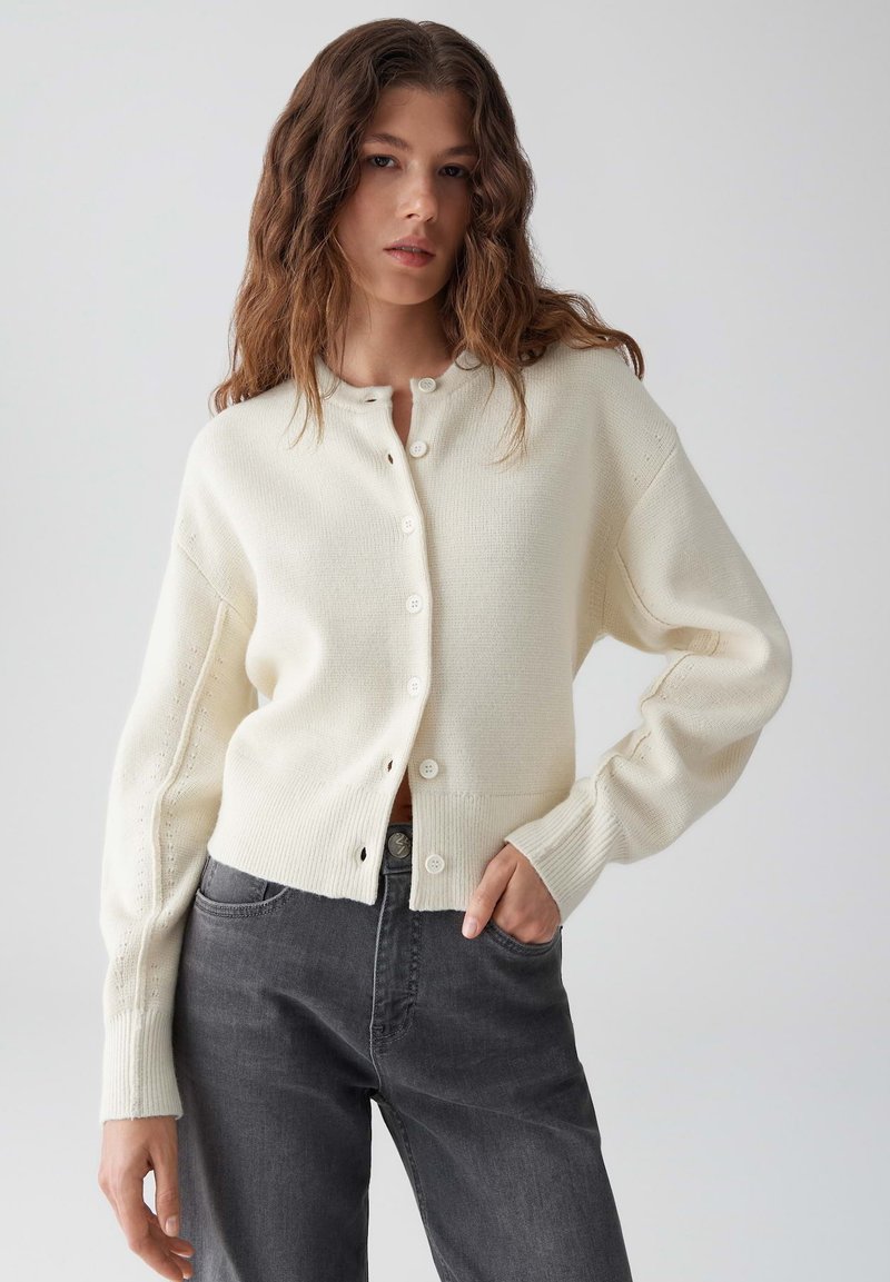 Witte gebreide cardigan met knopen aan de voorkant, verlaagde schouders en geribde manchetten; gedragen over donkergrijze jeans. Gladde textuur, casual ontwerp.