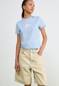 Ljusblå bomullströja med en rosa skalgrafik på bröstet; kombinerad med beige denimshorts som har en avslappnad passform och fickor.