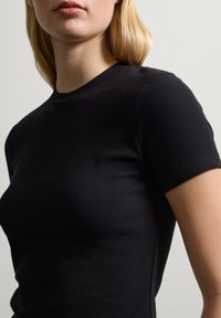 Blonde Person, der ein eng anliegendes schwarzes kurzärmliges Shirt aus Rippstoff trägt, von den Schultern bis zur Mitte des Oberkörpers vor einem schlichten Hintergrund gezeigt.