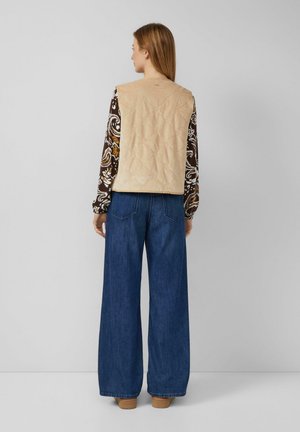 Vrouw met de rug naar de camera, gekleed in een beige gewatteerde vest, een patroon lange mouwen top, brede blauwe jeans en tan schoenen, tegen een effen achtergrond.