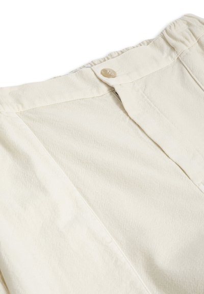 Gros plan sur la taille d'un pantalon couleur crème avec fermeture par bouton et détails de coutures visibles sur un tissu texturé.