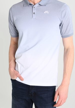 Mand iført en kortærmet grå Nike polo og mørkeblå jeans, står med den ene hånd i lommen.