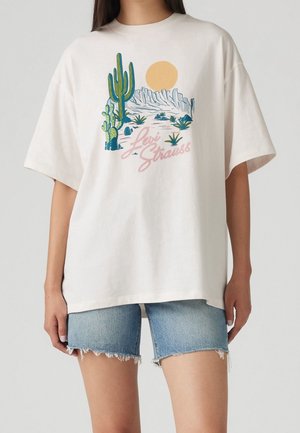 Vit oversize t-shirt med en grafisk bild av en kaktus och ett berg med en sol, kombinerad med ljusblå jeansshorts med fransad kant.