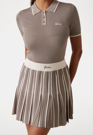 Kobieta w taupe dzianinowej koszulce polo z białymi lamówkami i dopasowanej plisowanej spódnicy z białymi pionowymi paskami i logo Guess na piersi i w pasie.