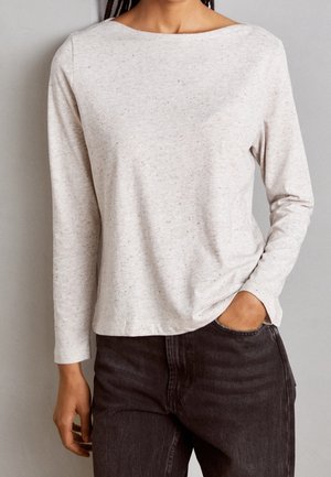 Kvinde iført en lys beige langærmet top med prikmønster og højtaljede mørkegrå jeans, den ene hånd i lommen.