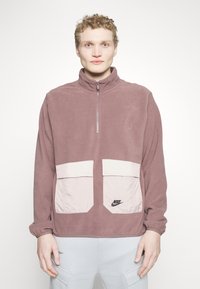 Mauve fleecová mikina na zips v štvrť dĺžke, so širokými krémovými prednými vreckami a čiernym logom Nike na spodnej ľavej strane.