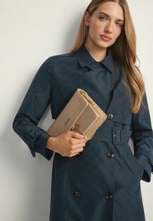 Kobieta w ciemnoniebieskim trenczu z paskiem, trzymająca beżową designerską kopertówkę z złotym łańcuszkiem na jasnym tle.