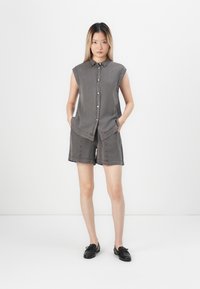 Ung kvinne står i en ermeløs grå skjorte med knapper og matchende shorts, iført svarte slip-on sko, med hendene i lommene, mot en nøytral bakgrunn.