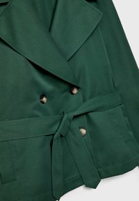 Manteau vert foncé ceinturé avec larges revers et double boutonnage, montrant des poches avant et des coutures détaillées.