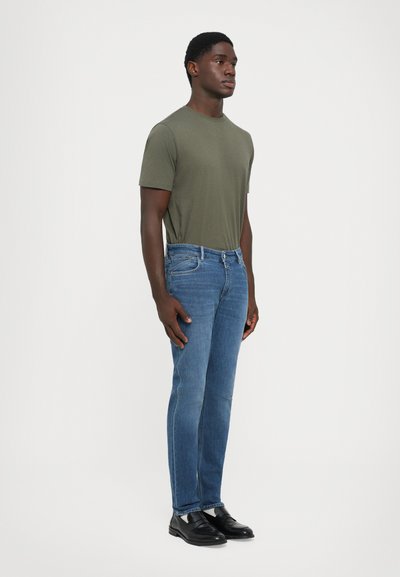 Man stands facing sideways wearing olive green short-sleeve shirt, blue jeans, white socks, and black leather shoes on plain light background.