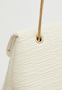 Biała torebka o teksturowanej powierzchni z zaokrąglonym dnem, z złotą plecioną łańcuszkową rączką i okrągłym detalem z grometem.