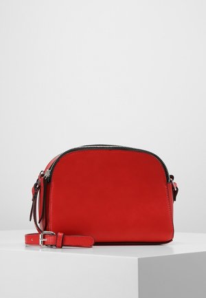 Czerwona skórzana torba na ramię z czarnym zamkiem błyskawicznym, regulowanym paskiem i srebrną klamrą spoczywająca na białym piedestale na szarym tle.