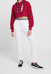 Hoodie rossa cropped con lacci neri e bianchi, abbinato a joggers bianchi con vita a scacchi e sneakers nere.