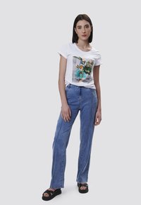 Camiseta blanca de algodón con un estampado gráfico, combinada con pantalones de mezclilla azul claro con piernas rectas y detalles en la costura lateral. Sandalias negras.