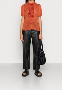 Blusa rossa con motivo a fantasia e dettaglio arricciato al colletto, abbinata a pantaloni in pelle nera e una borsa in tessuto nera. Indossate sandali neri spessi.