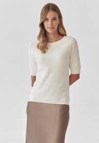 Strik cremefarvet sweater med ribbet halsudskæring og korte ærmer, parret med en glat, lysebrun nederdel. Enkel, moderne design med en afslappet pasform.