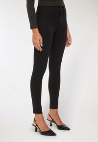Persona che indossa jeans skinny neri, tacchi slingback a punta nera e un maglione lungo a coste scuro, in piedi contro uno sfondo bianco.