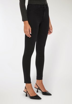 Persona che indossa jeans skinny neri, tacchi slingback a punta nera e un maglione lungo a coste scuro, in piedi contro uno sfondo bianco.