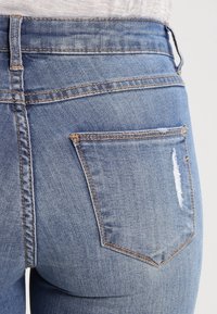 Vaqueros de mezclilla azul claro con cintura alta, que presentan un sutil desgastado, un bolsillo trasero rasgado y costuras en contraste.