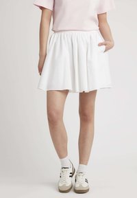 Jupe évasée blanche avec une taille élastique et une poche latérale. Portée avec un haut rose et des baskets, sur un fond uni.