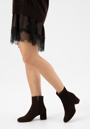 Žena v černých kotníkových botách s blokovým podpatkem a černé sukni s krajkovým lemem, kráčející na jednobarevném bílém pozadí.