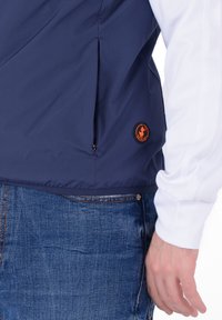 Giacca a vento blu navy con tasca con zip e logo arancione. La manica è bianca, caratterizzata da una texture liscia e un design sportivo.