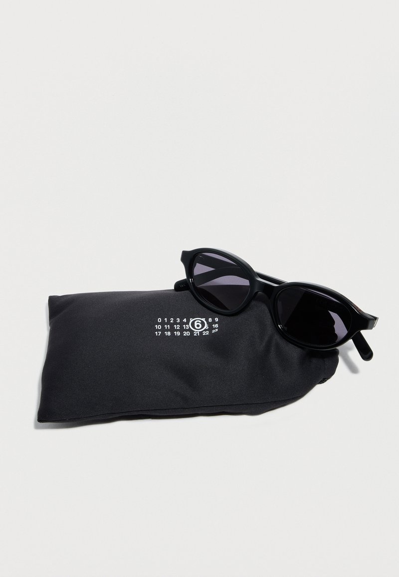 Gafas de sol ovaladas negras descansando sobre una funda blanda negra con un diseño numérico blanco sobre un fondo claro.