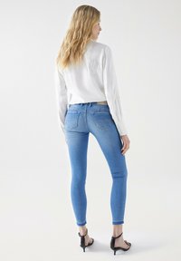 Blusa blanca con bordado, combinada con jeans ajustados azul con cintura media y dos bolsillos traseros. Sandalias de tacón negras.