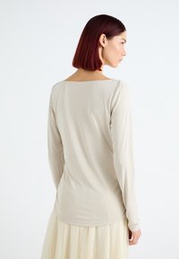 Langermet, beige topp med vid utringning og figurtilpasset silhuett, laget av glatt stoff. Brukt med en flytende, lys farget skjørt.