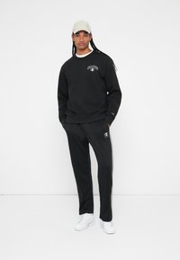 Sweatshirt preto e calças de fato desportivo com um logótipo. O sweatshirt tem um decote arredondado, enquanto as calças têm riscas laterais. Combinado com ténis brancos e um boné.