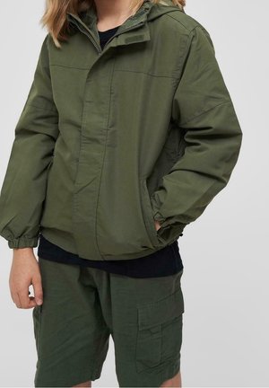 Olivgrüne Jacke mit Kapuze, elastischen Bündchen und Seitentaschen. Getragen über einem schwarzen Shirt und kombiniert mit grünen Cargo-Shorts.