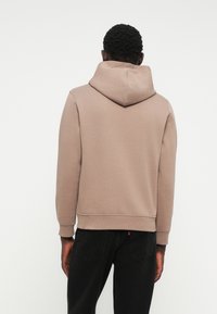 Felpa con cappuccio marrone realizzata in tessuto morbido. Presenta una tasca a marsupio, polsini a coste e una vestibilità rilassata. Appare indossata con jeans neri.