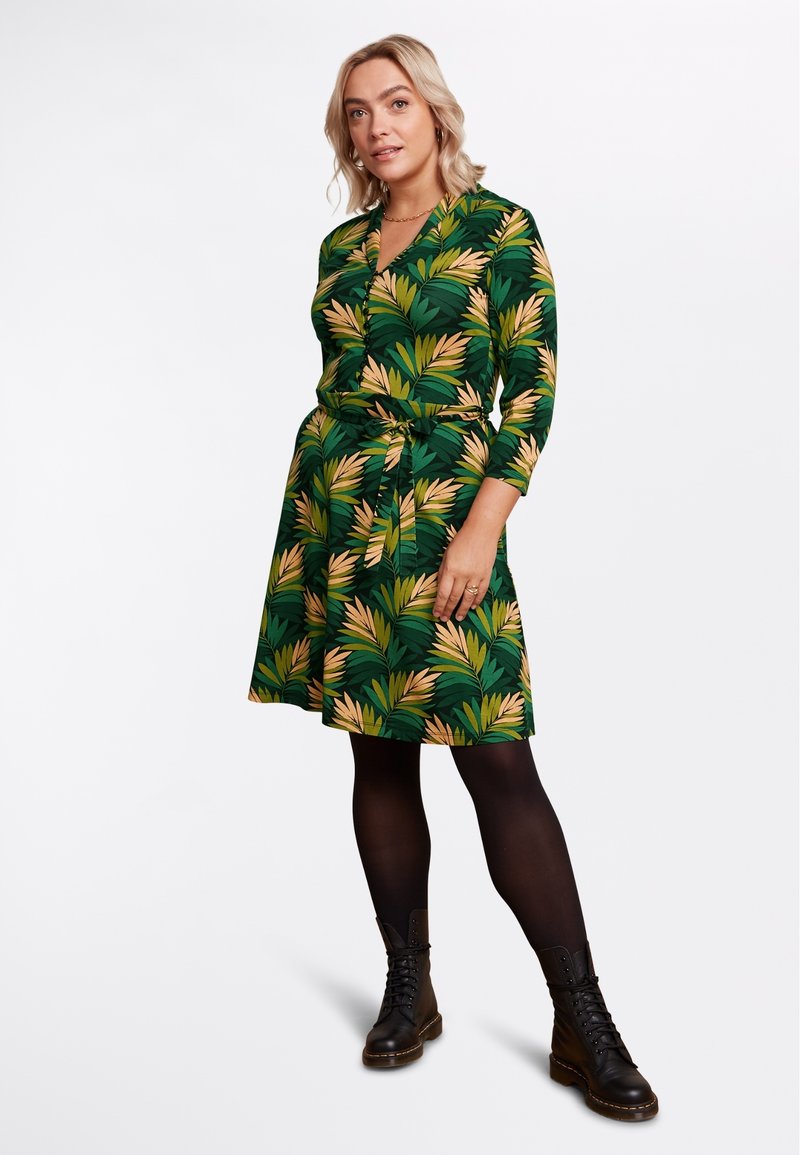 Abito verde con un motivo a foglia, maniche a tre quarti, vita cinchata e gonna svolazzante. Abbinato a stivali militari neri.