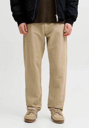 Homme portant un pantalon beige droit, des mocassins lacés couleur sable, un pull marron foncé et une veste noire zippée, debout devant un fond blanc.