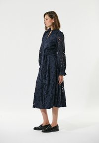 Marineblaues Spitzenkleid mit floralen Mustern, langen Ärmeln, knöpfbarem Kragen, tailliertem Bund und Rock in Midi-Länge. Kombiniert mit schwarzen Loafers.