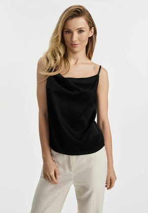 Czarna satynowa koszulka na ramiączkach z drapowanym dekoltem i cienkimi ramiączkami, zestawiona z jasnobeżowymi spodniami, podkreślająca miękką, płynącą sylwetkę.