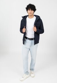 Navy Zip-up-Jacke mit Kapuze, mit einem gesteppte Design, kombiniert mit einem weißen T-Shirt, hellblauen Jeans und weißen Sneakern.