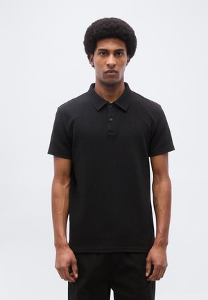 Giovane uomo con acconciatura afro che indossa una polo nera a maniche corte e pantaloni neri, in piedi contro uno sfondo chiaro e uniforme.