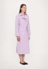 Lilac krāsu iešūts mētelis ar dubultā pogu dizainu, platām atloks un jostu viduklī. Garas piedurknes un vidus teļu garums. Melni apavi.