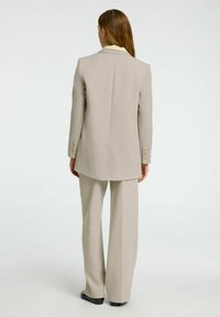 Traje ajustado de un beige claro que presenta un blazer largo con solapas en pico y pantalones de pierna ancha. Textura sutil con detalles de botones en las mangas.