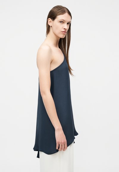 Hosszú barna hajú nő ujjatlan, aszimmetrikus sötétkék felsőben és fehér nadrágban, világos egyszínű háttér előtt áll.